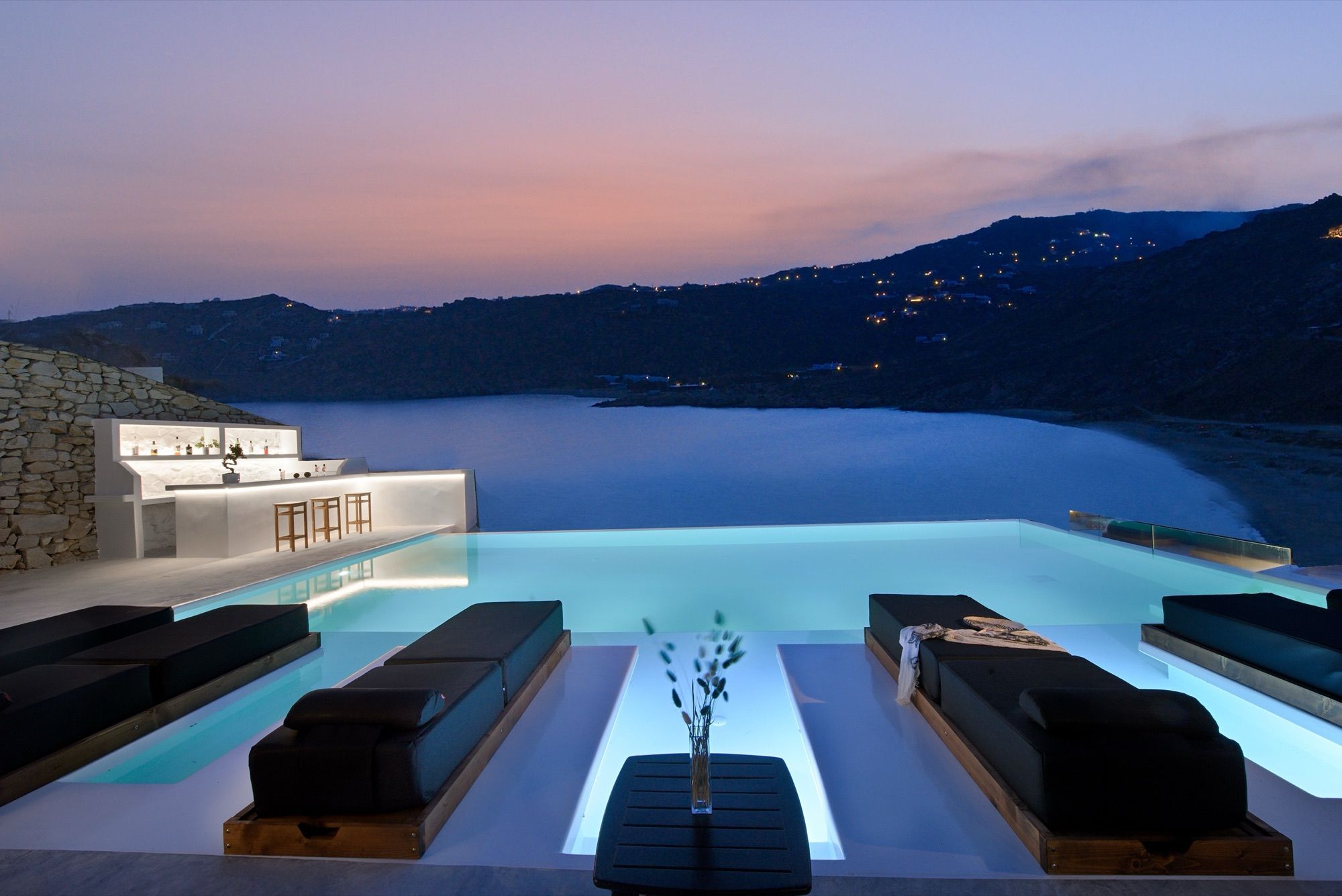Infinity pool with in-water sunbeds and the pool bar at dusk, Elia bay and lit hillside behind