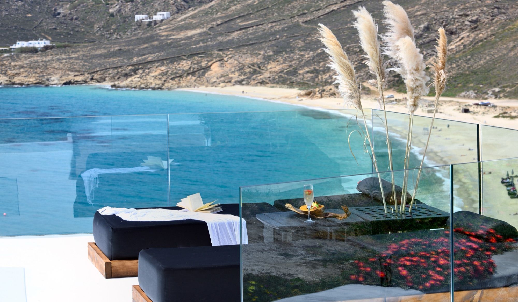 Infinity pool edge with sunbeds, pampas grass and a glass balustrade above the turquoise bay of Elia Beach