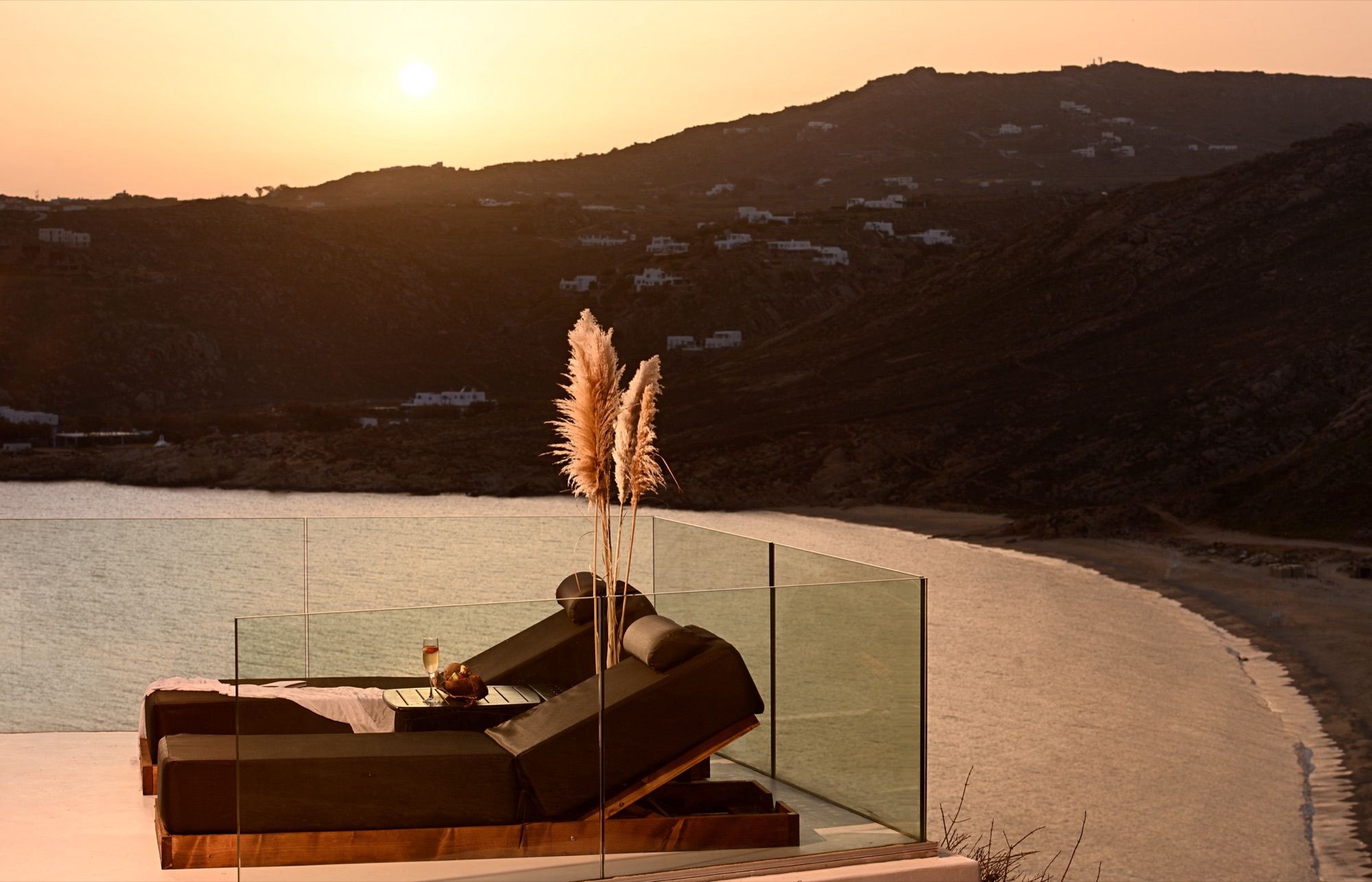 Double sunbed beside pampas grass and a glass balustrade at sunset over Elia Beach
