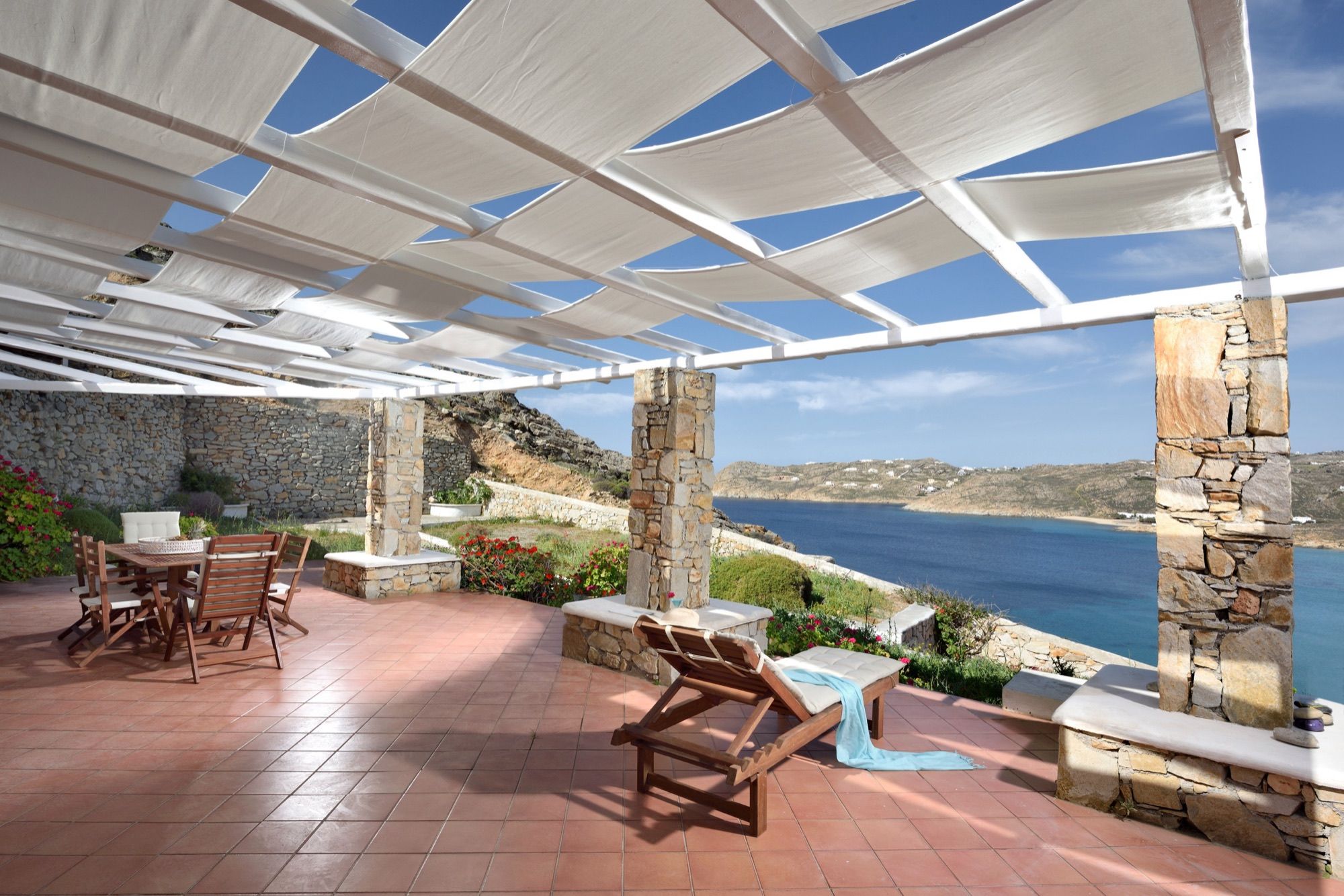 Terrace under a white pergola with stone pillars, a lounger and a dining table, looking over the bay