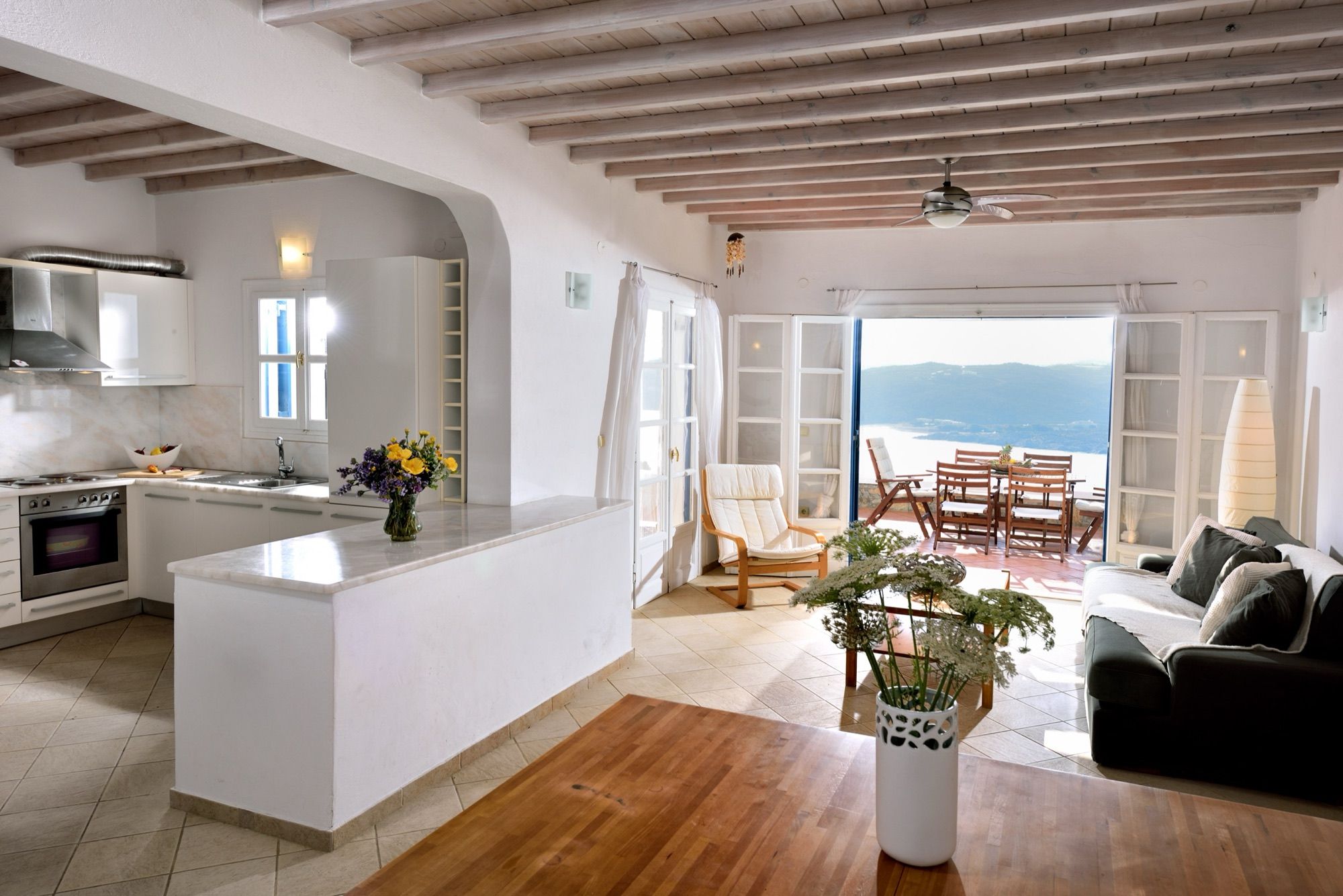Open-plan living room and kitchen under a beamed ceiling, with doors to the terrace