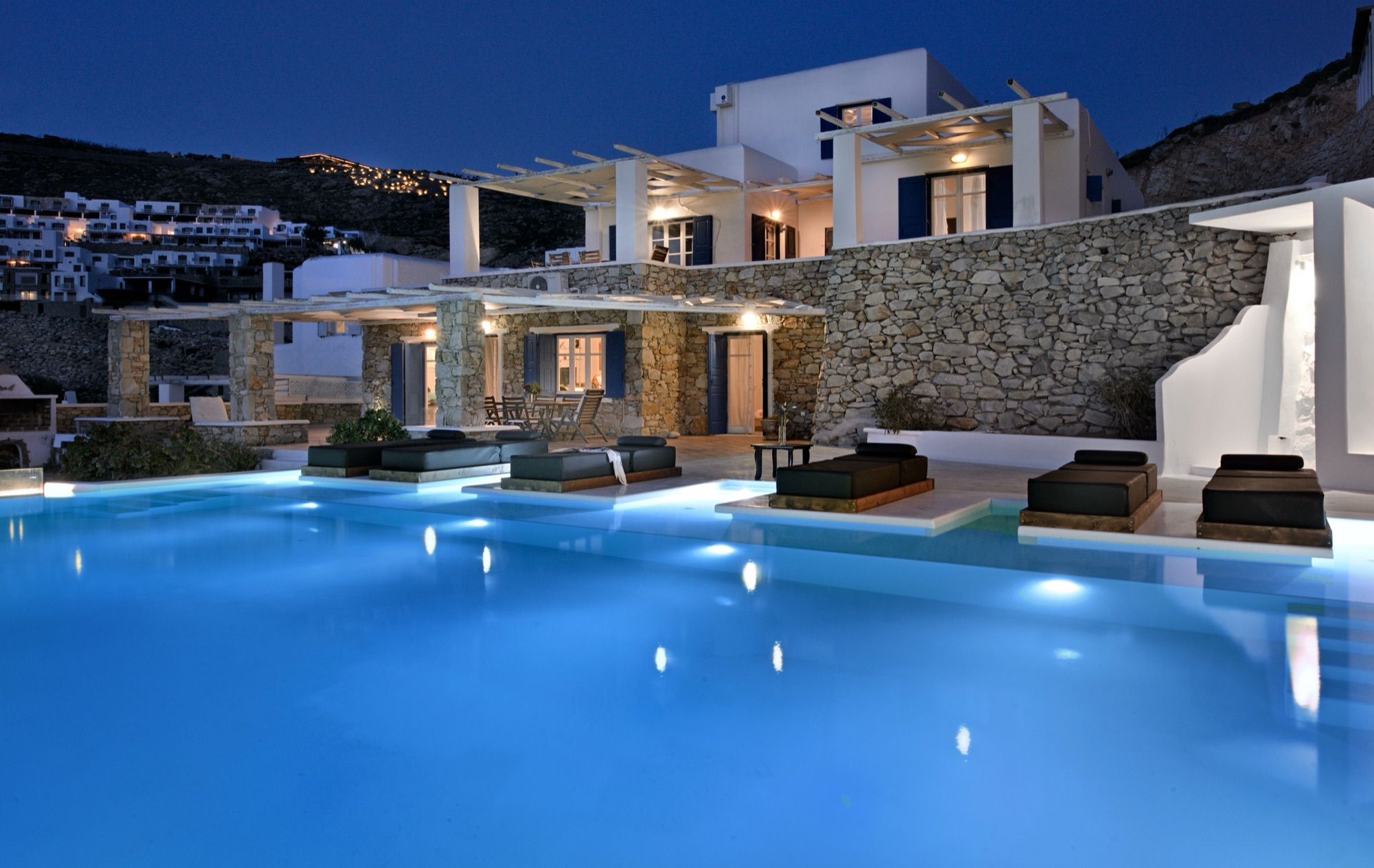 Villa Chrysta lit up at dusk above its pool, with stone walls, pergolas and sunbeds